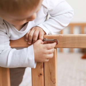 May include: A light-colored wooden crib with a metal latch. A child in a white long-sleeved shirt is shown interacting with the latch. The crib's wooden frame is visible, showcasing its construction and design.