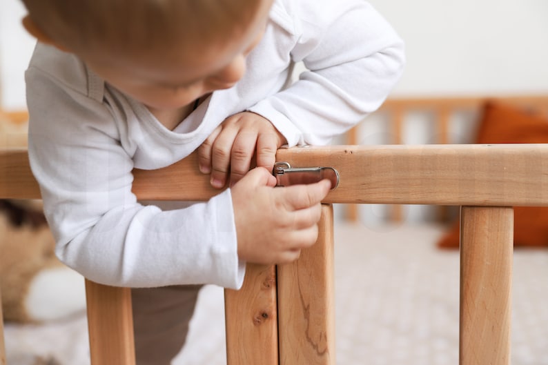May include: A light-colored wooden crib with a metal latch. A child in a white long-sleeved shirt is shown interacting with the latch. The crib's wooden frame is visible, showcasing its construction and design.