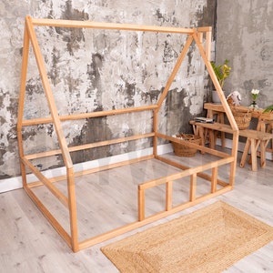 May include: A natural wood-framed house-shaped bed frame. The bed frame is on a light wood floor with a woven jute rug. A white storage box with a bunny face is on the left. A small wooden table and benches are in the background.