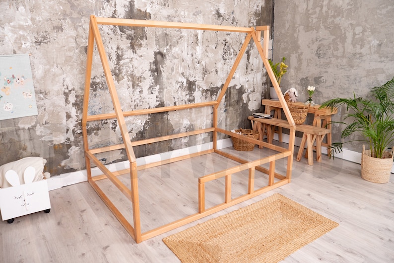 May include: A natural wood-framed house-shaped bed frame. The bed frame is on a light wood floor with a woven jute rug. A white storage box with a bunny face is on the left. A small wooden table and benches are in the background.