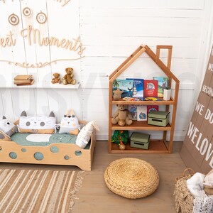 May include: A light wood children's bed with a green mattress and cat-shaped pillows. A wooden bookshelf with a house-shaped top is filled with books and toys. A woven rug and a round woven ottoman are on the floor. A sign with the text "WE DO FUN WE LOVE" is on the wall.