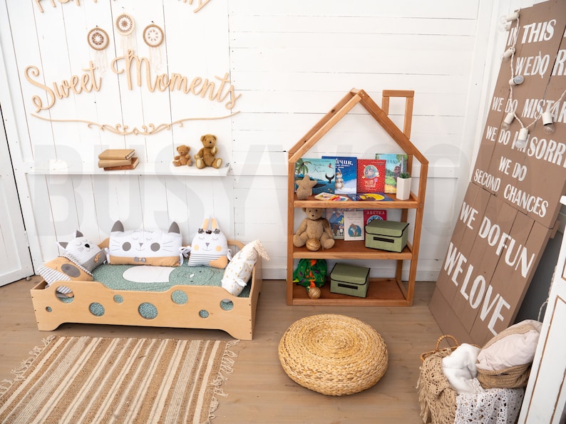 May include: A light wood children's bed with a green mattress and cat-shaped pillows. A wooden bookshelf with a house-shaped top is filled with books and toys. A woven rug and a round woven ottoman are on the floor. A sign with the text "WE DO FUN WE LOVE" is on the wall.