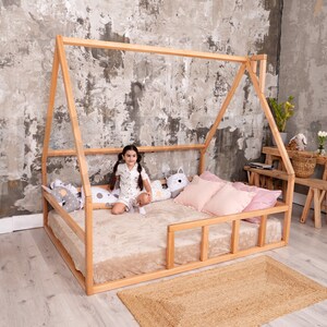 May include: A wooden house-shaped bed frame with a beige mattress, pink pillows, and decorative pillows. A small girl sits on the bed. A woven rug is on the floor. The room has a neutral color palette.