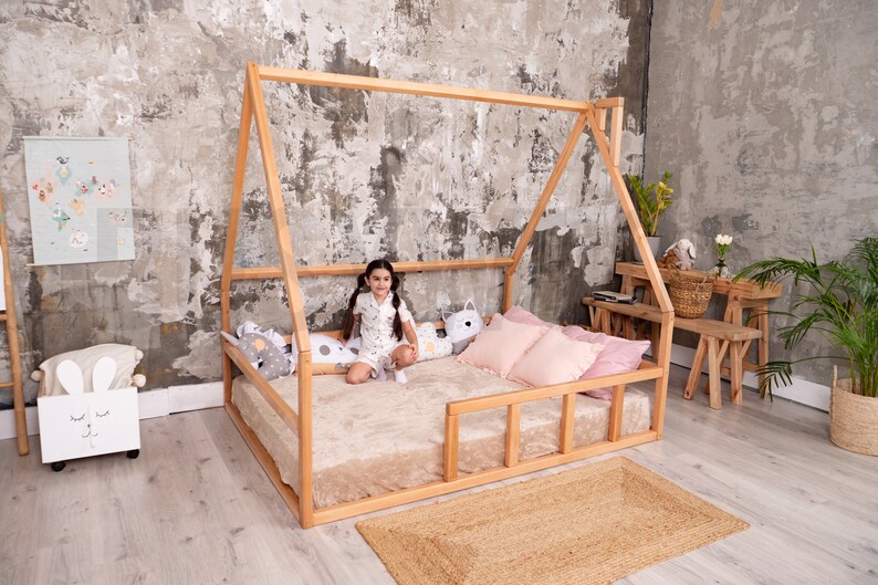May include: A wooden house-shaped bed frame with a beige mattress, pink pillows, and decorative pillows. A small girl sits on the bed. A woven rug is on the floor. The room has a neutral color palette.