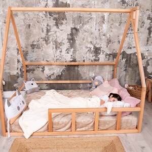May include: A wooden house-shaped bed frame with a mattress, pillows, and a blanket. The bed is in a room with a gray and white textured wall. Decorative pillows and stuffed animals are on the bed.