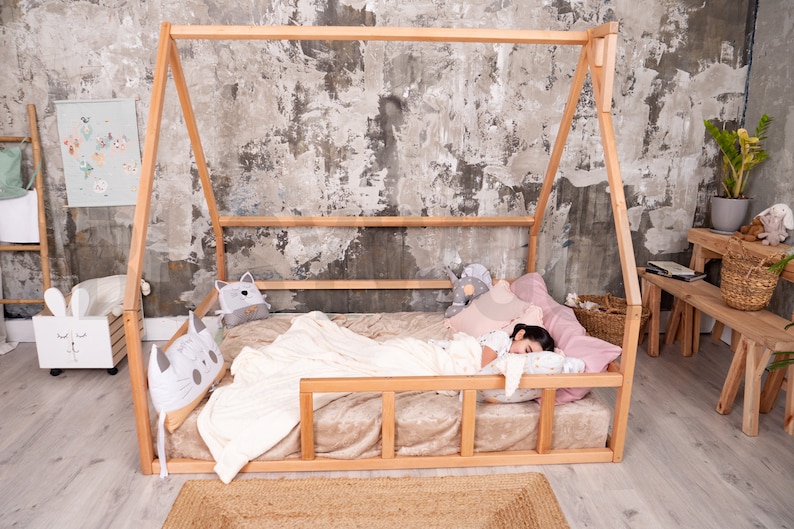 May include: A wooden house-shaped bed frame with a mattress, pillows, and a blanket. The bed is in a room with a gray and white textured wall. Decorative pillows and stuffed animals are on the bed.