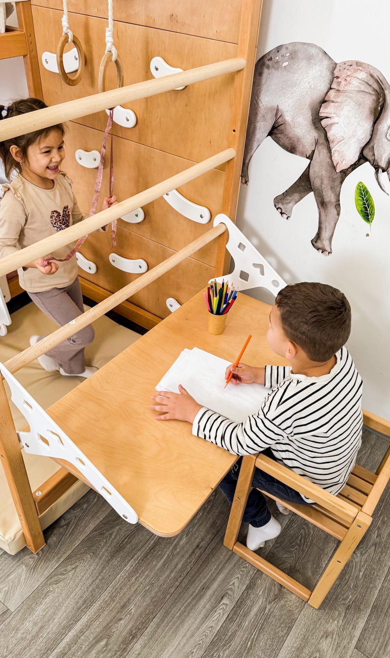 Pu&ograve; includere: Una struttura per arrampicata in legno con una scrivania e una sedia. Un bambino disegna alla scrivania con una matita. Un altro bambino gioca sulla struttura. &Egrave; visibile un adesivo da parete a forma di elefante.