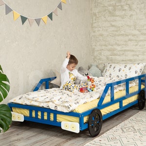 K&ouml;nnte beinhalten: Ein blaues Holzbett in Form eines Autos mit schwarzen R&auml;dern und gelben Scheinwerfern. Ein Kind sitzt auf dem Bett und spielt mit Spielzeug.