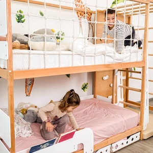 Pu&ograve; includere: Un letto a castello in legno con struttura marrone chiaro, dettagli bianchi e una rete di corda. Un bambino sta salendo sul letto superiore. Il letto inferiore ha un lenzuolo rosa. Il muro ha decalcomanie di giraffe e foglie.