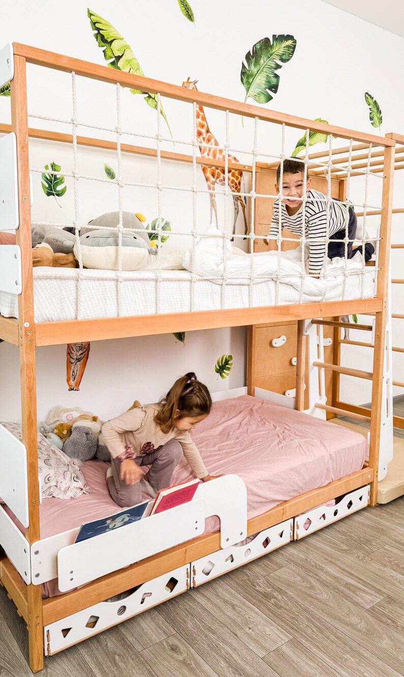 Pu&ograve; includere: Un letto a castello in legno con struttura marrone chiaro, dettagli bianchi e una rete di corda. Un bambino sta salendo sul letto superiore. Il letto inferiore ha un lenzuolo rosa. Il muro ha decalcomanie di giraffe e foglie.