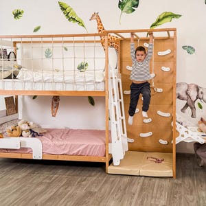 Pu&ograve; includere: Letto a castello in legno con parete da arrampicata, scala e sbarre per scimmie. Il letto ha una sponda di sicurezza bianca e una trapunta rosa sulla cuccetta inferiore. Un bambino &egrave; appeso alle sbarre per scimmie. Foglie decorative e una giraffa sul muro.