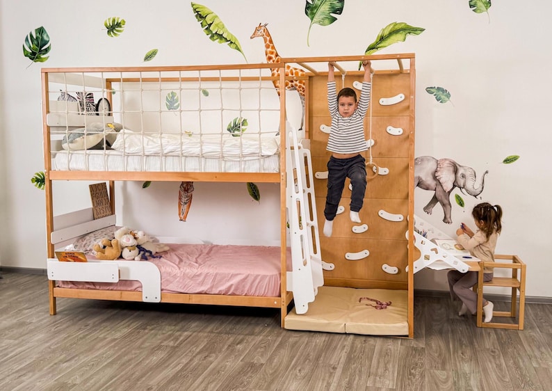 Pu&ograve; includere: Letto a castello in legno con parete da arrampicata, scala e sbarre per scimmie. Il letto ha una sponda di sicurezza bianca e una trapunta rosa sulla cuccetta inferiore. Un bambino &egrave; appeso alle sbarre per scimmie. Foglie decorative e una giraffa sul muro.