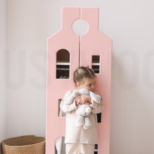 Op de afbeelding: Een roze houten speelhuis met een witte deur en een klein kind dat een knuffeldier vasthoudt. Het huis heeft planken en een hangende kledingrek.