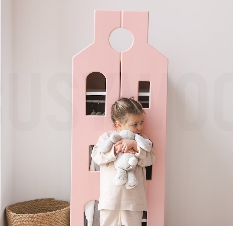 Op de afbeelding: Een roze houten speelhuis met een witte deur en een klein kind dat een knuffeldier vasthoudt. Het huis heeft planken en een hangende kledingrek.