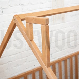 May include: Close-up of a wooden bed frame with a house-shaped design. The frame is made of light-colored wood and features a slatted headboard and footboard. The structure is assembled with visible joints and screws.