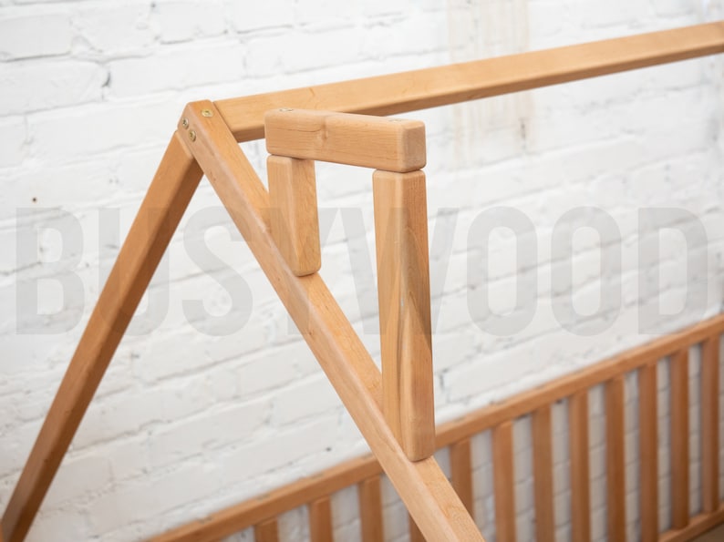 May include: Close-up of a wooden bed frame with a house-shaped design. The frame is made of light-colored wood and features a slatted headboard and footboard. The structure is assembled with visible joints and screws.