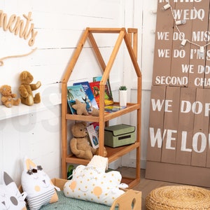 May include: A wooden bookshelf shaped like a house with three shelves. The shelves are filled with books and stuffed animals. The bookshelf is in a room with white walls and a wooden sign with the text "We do real, We do mistakes, We do I'm sorry, We do second chances, We do fun, We love".