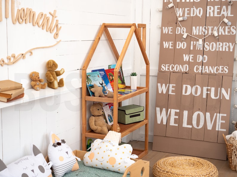 May include: A wooden bookshelf shaped like a house with three shelves. The shelves are filled with books and stuffed animals. The bookshelf is in a room with white walls and a wooden sign with the text "We do real, We do mistakes, We do I'm sorry, We do second chances, We do fun, We love".
