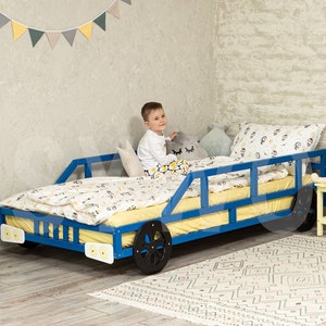 Op de afbeelding: Een blauw houten autobed met een gele matras en witte beddengoed. Een kind zit op het bed. Het bed heeft zwarte wielen en witte koplampen.