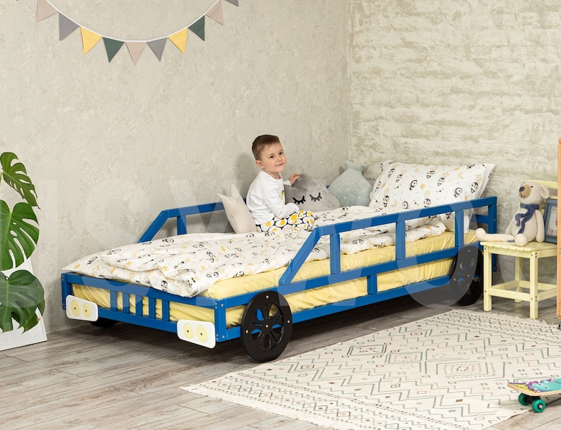 K&ouml;nnte beinhalten: Ein blaues Holzbett in Form eines Autos mit einer gelben Matratze und wei&szlig;en Bettw&auml;sche. Ein Kind sitzt auf dem Bett. Das Bett hat schwarze R&auml;der und wei&szlig;e Scheinwerfer.