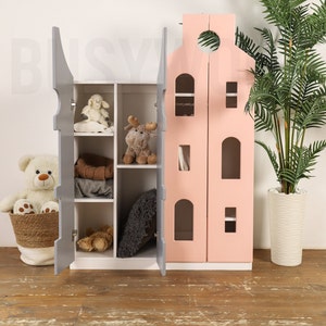 Op de afbeelding: Een grijs en roze houten speelhuis met een deur die open kan. Het huis heeft planken binnenin en is versierd met ramen en een dak. Het huis staat op een houten vloer.