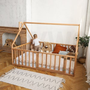 May include: A wooden house-shaped bed frame with a slatted side and gate. The bed is filled with a mattress and pillows. A child is standing inside the bed with a teddy bear. A rug with a geometric pattern is on the floor.