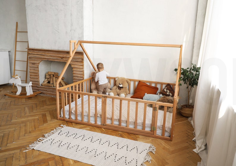 May include: A wooden house-shaped bed frame with a slatted side and gate. The bed is filled with a mattress and pillows. A child is standing inside the bed with a teddy bear. A rug with a geometric pattern is on the floor.