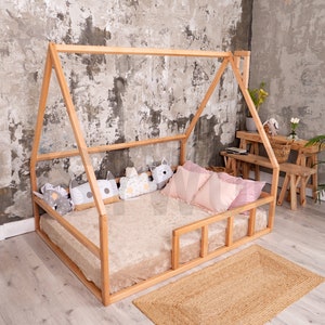 May include: A natural wood house-shaped bed frame with a plush beige mattress and decorative pillows. The bed is on a woven jute rug. The room has a neutral color palette with a concrete-style wall.