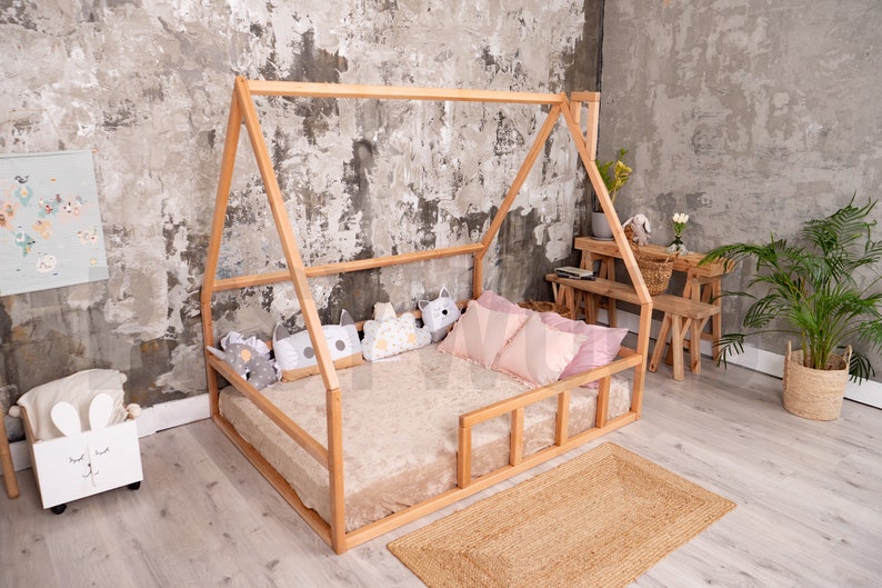 May include: A natural wood house-shaped bed frame with a plush beige mattress and decorative pillows. The bed is on a woven jute rug. The room has a neutral color palette with a concrete-style wall.