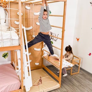 Pu&ograve; includere: Una struttura da gioco in legno per bambini con parete da arrampicata, scala e sbarre. Un bambino con una maglietta a righe &egrave; appeso alle sbarre. Un altro bambino &egrave; seduto a una piccola scrivania. La struttura comprende un letto e un materassino.