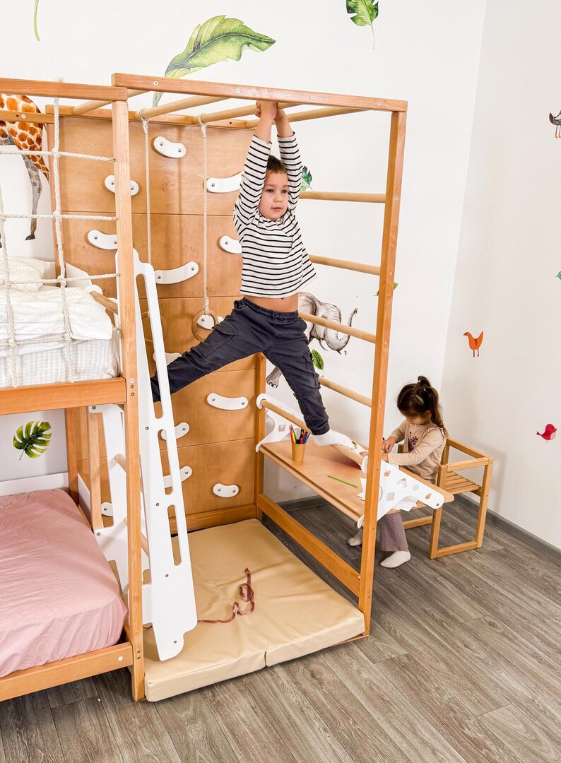 Pu&ograve; includere: Una struttura da gioco in legno per bambini con parete da arrampicata, scala e sbarre. Un bambino con una maglietta a righe &egrave; appeso alle sbarre. Un altro bambino &egrave; seduto a una piccola scrivania. La struttura comprende un letto e un materassino.