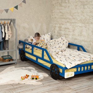 K&ouml;nnte beinhalten: Ein blaues Holzbett in Form eines Autos mit schwarzen R&auml;dern und wei&szlig;en Scheinwerfern. Ein Kind sitzt auf dem Bett und liest ein Buch. Das Bett hat eine gelbe Matratze und eine Bettw&auml;sche mit wei&szlig;en und schwarzen Mustern. Das Bett steht in einem Zimmer mit einer grauen Wand und einem Holzkleiderst&auml;nder.