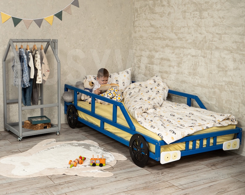 K&ouml;nnte beinhalten: Ein blaues Holzbett in Form eines Autos mit schwarzen R&auml;dern und wei&szlig;en Scheinwerfern. Ein Kind sitzt auf dem Bett und liest ein Buch. Das Bett hat eine gelbe Matratze und eine Bettw&auml;sche mit wei&szlig;en und schwarzen Mustern. Das Bett steht in einem Zimmer mit einer grauen Wand und einem Holzkleiderst&auml;nder.