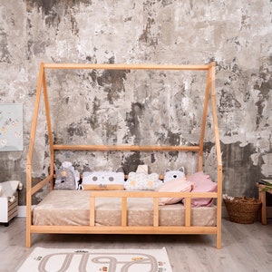 May include: A wooden teepee bed frame with a beige mattress and pillows. The bed is in a room with a concrete wall and a wooden ladder with a green bag hanging on it.