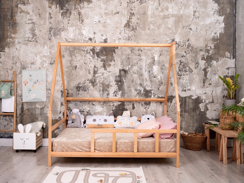 May include: A wooden teepee bed frame with a beige mattress and pillows. The bed is in a room with a concrete wall and a wooden ladder with a green bag hanging on it.