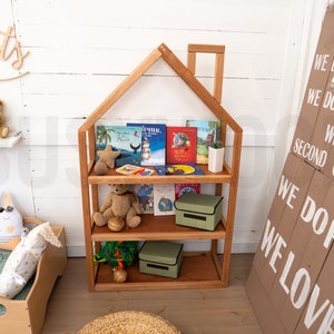 May include: A wooden bookshelf shaped like a house with three shelves. The shelves are filled with children's books and stuffed animals. There are two green storage bins on the bottom shelves. The bookshelf is in a room with white walls and a wooden floor.