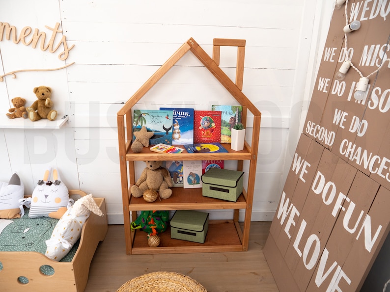 May include: A wooden bookshelf shaped like a house with three shelves. The shelves are filled with children's books and stuffed animals. There are two green storage bins on the bottom shelves. The bookshelf is in a room with white walls and a wooden floor.