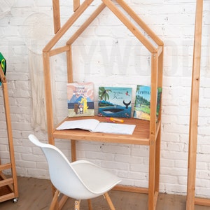 K&ouml;nnte beinhalten: Ein h&ouml;lzerner Spielhaus-Schreibtisch mit einem wei&szlig;en Stuhl und einem braunen Teppich. Der Schreibtisch hat ein Regal mit drei B&uuml;chern darauf. Die B&uuml;cher sind betitelt mit "Теплая Свитер для Альмута", "Грана в Ким", und "Гургурал".