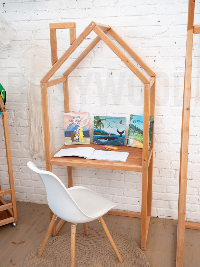 K&ouml;nnte beinhalten: Ein h&ouml;lzerner Spielhaus-Schreibtisch mit einem wei&szlig;en Stuhl und einem braunen Teppich. Der Schreibtisch hat ein Regal mit drei B&uuml;chern darauf. Die B&uuml;cher sind betitelt mit "Теплая Свитер для Альмута", "Грана в Ким", und "Гургурал".