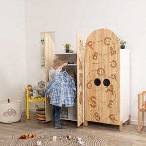 May include: A wooden children's wardrobe with two doors featuring a white background and a brown wood grain pattern. The doors are decorated with a whimsical design of letters and circles. A young child is standing in front of the wardrobe, holding a blue and white plaid shirt.