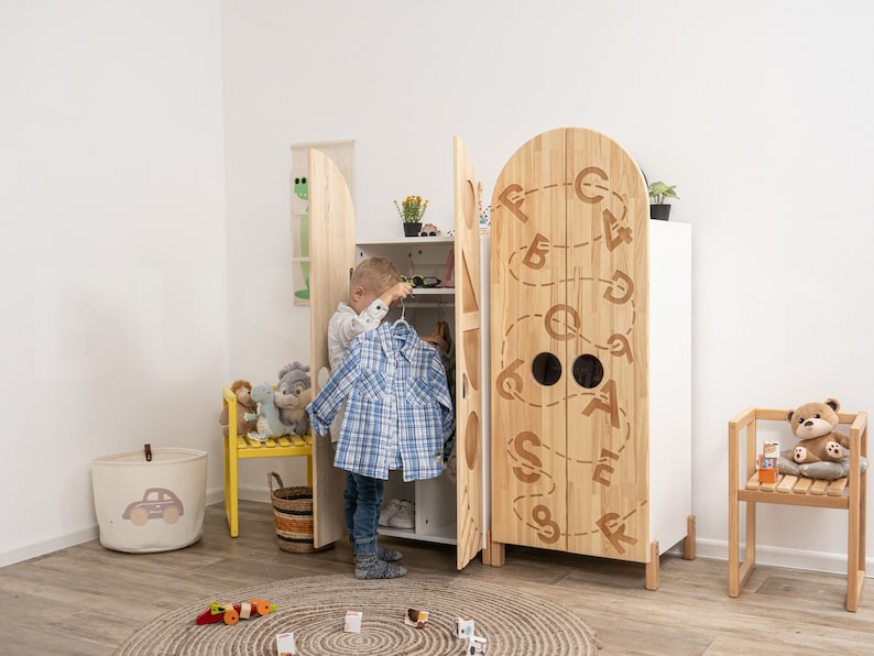 May include: A wooden children's wardrobe with two doors featuring a white background and a brown wood grain pattern. The doors are decorated with a whimsical design of letters and circles. A young child is standing in front of the wardrobe, holding a blue and white plaid shirt.