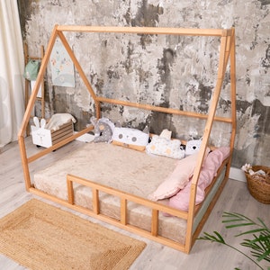 May include: A wooden house-shaped bed frame with a beige mattress and pillows. The bed is on a jute rug. Decorative pillows in various shapes and colors are arranged on the bed. The room has a neutral color palette with a textured wall.