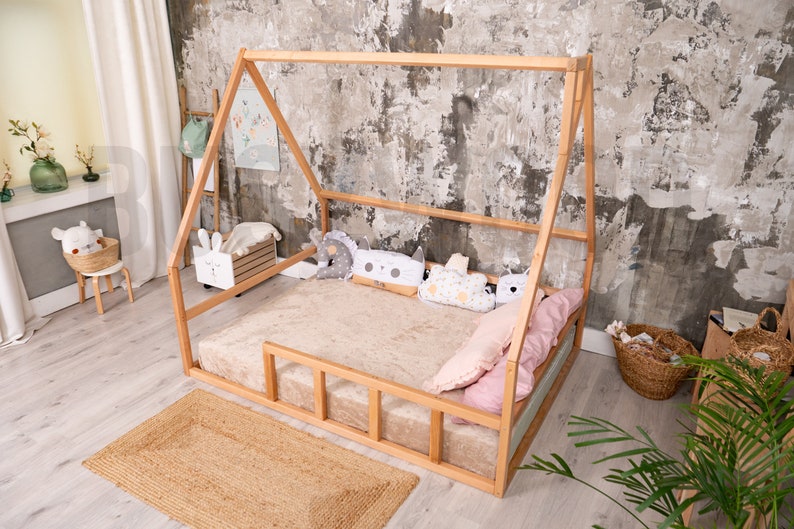 May include: A wooden house-shaped bed frame with a beige mattress and pillows. The bed is on a jute rug. Decorative pillows in various shapes and colors are arranged on the bed. The room has a neutral color palette with a textured wall.