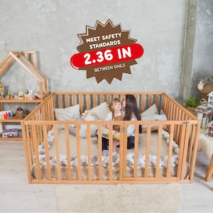 Op de afbeelding: Een lichtbruine houten box met verticale spijlen en een zacht, lichtgekleurd interieur. De box heeft een bord met de tekst "2.36 IN TUSSEN DE RELINGEN". Een kind en een volwassene zitten erin.