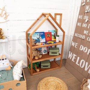 May include: A wooden bookshelf shaped like a house with three shelves. The shelves are filled with children's books and two green storage boxes. The bookshelf is in a room with a white wall and a wooden sign with the text "WE DO FUN WE LOVE" on it.