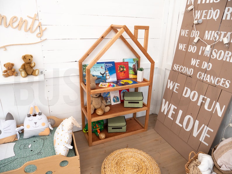 May include: A wooden bookshelf shaped like a house with three shelves. The shelves are filled with children's books and two green storage boxes. The bookshelf is in a room with a white wall and a wooden sign with the text "WE DO FUN WE LOVE" on it.