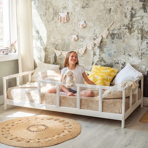 Op de afbeelding: Een wit houten peuterbed met een beige matras en kussens. Een jong persoon zit op het bed en houdt een knuffel vast. Een rond jute vloerkleed ligt op de vloer. De kamer heeft een neutraal kleurenpalet.