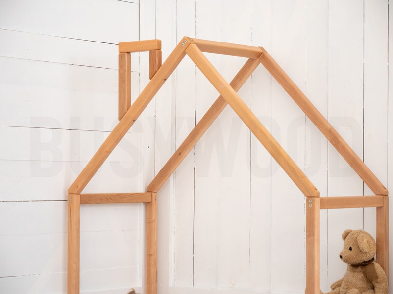 May include: A wooden playhouse frame with a simple design, made of natural wood. The frame is unfinished and ready to be painted or stained. A teddy bear sits inside the frame.