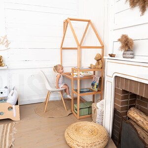 May include: A white room with a wooden playhouse desk and chair. A young girl is sitting at the desk. There are books on the shelves of the playhouse. There is a fireplace with a brick surround and a wooden mantle. There is a rug on the floor and a stuffed animal on the shelf.