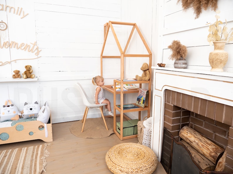 May include: A white room with a wooden playhouse desk and chair. A young girl is sitting at the desk. There are books on the shelves of the playhouse. There is a fireplace with a brick surround and a wooden mantle. There is a rug on the floor and a stuffed animal on the shelf.